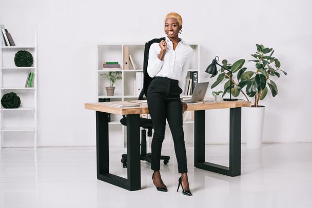 cheerful african american businesswoman with short hair holding jacket in modern office - Fotó, kép