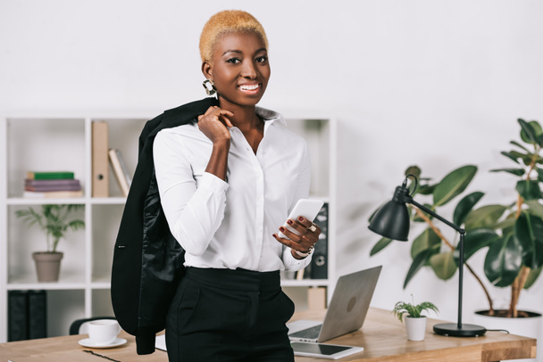 african american businesswoman with short hair holding smartphone and jacket in modern office - Fotografie, Obrázek