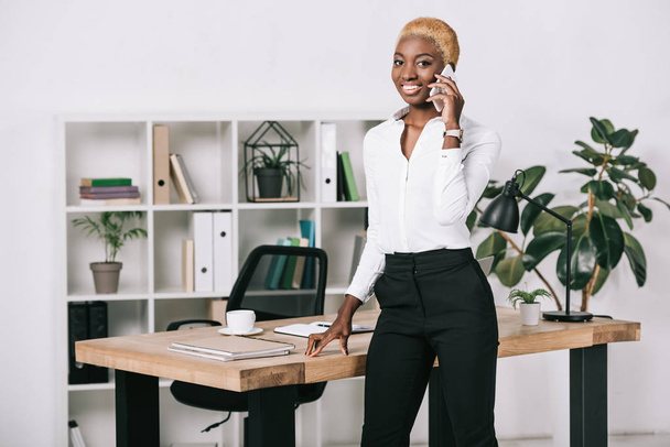 atraktivní africká americká podnikatelka s krátkými vlasy na smartphone v moderní kanceláři - Fotografie, Obrázek