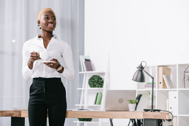 cheerful african american businesswoman with short hair holding cup with drink in modern office  - Foto, Bild