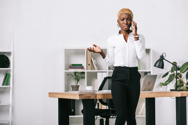 upset african american businesswoman with short hair talking on smartphone in modern office - Фото, зображення