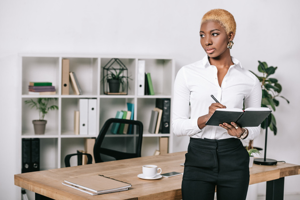 zamyšlený africká americká podnikatelka s krátkými vlasy, stojící vedle stolu a psaní v poznámkovém bloku  - Fotografie, Obrázek