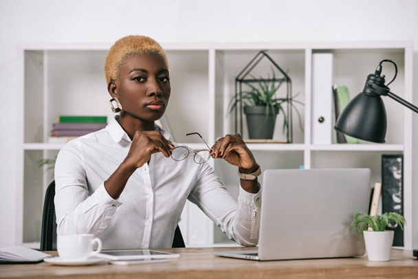 уверенная африканская американская предпринимательница с короткими волосами, держащая очки в современном офисе
 - Фото, изображение