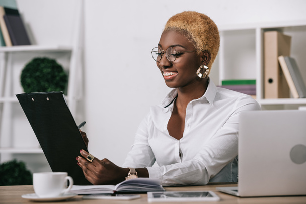 alegre afroamericana mujer de negocios sosteniendo portapapeles en oficina moderna
 - Foto, imagen
