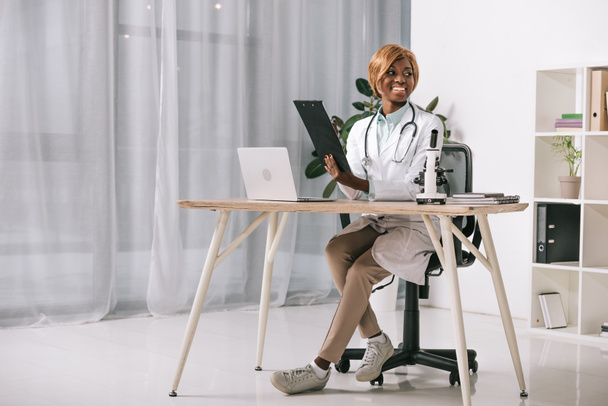atractivo científico afroamericano sentado con estetoscopio y sujetando el portapapeles
 - Foto, Imagen