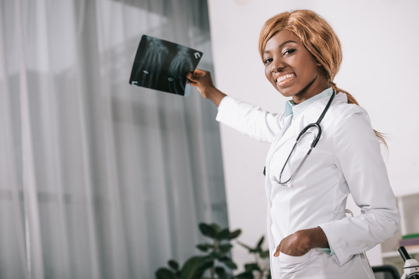 χαμογελώντας αφρικανική αμερικανική γυναίκα γιατρό εκμετάλλευση ακτινογραφία στην κλινική ενώ βλέπουν τα φωτογραφικών μηχανών - Φωτογραφία, εικόνα