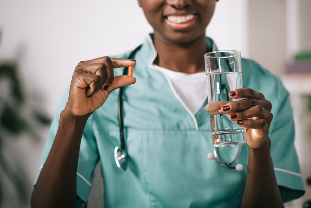 обрезанный вид улыбающейся африканской американской медсестры со стетоскопом, держащей таблетки и стакан воды
 - Фото, изображение