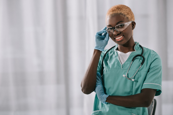 χαμογελαστά αφρικανική αμερικανική νοσοκόμα σε γάντια κρατώντας γυαλιά ενώ κοιτάζοντας τα φωτογραφικών μηχανών - Φωτογραφία, εικόνα