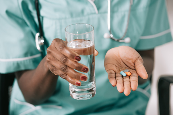 cropped view of african american nurse with stethoscope holding glass of water and pills in hand - Foto, Bild
