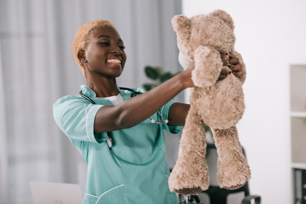 χαμογελώντας αφρικανική αμερικανική νοσοκόμα με στηθοσκόπιο κρατώντας αρκουδάκι  - Φωτογραφία, εικόνα