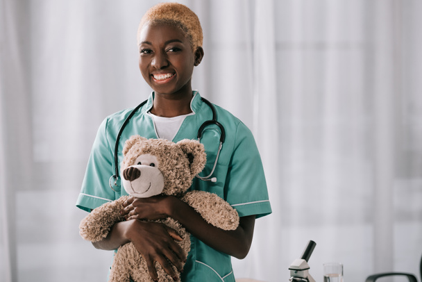 vrolijke Afrikaanse Amerikaanse verpleegster teddybeer houden in lichte kliniek - Foto, afbeelding