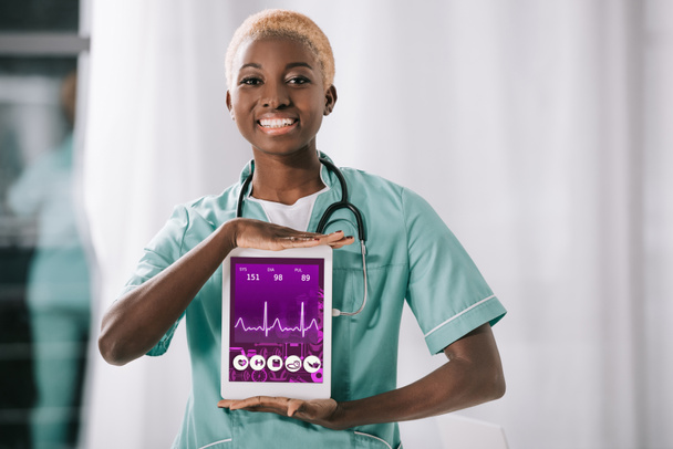 vrolijke Afrikaanse Amerikaanse verpleegster met stethoscoop holding digitale tablet met hartslag tarief op scherm - Foto, afbeelding