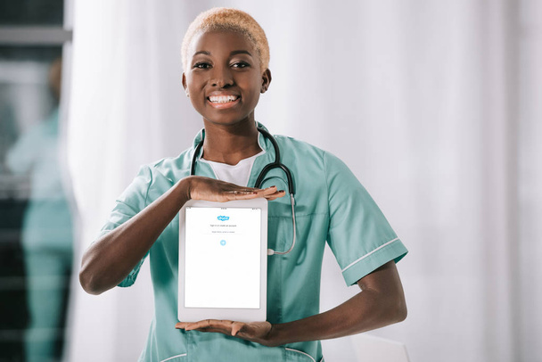 attractive cheerful african american nurse with stethoscope holding digital tablet with skype app - Zdjęcie, obraz
