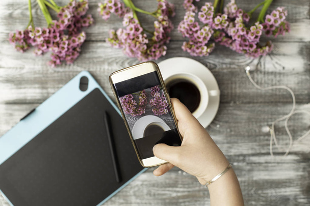 Instagram φωτογράφος blogging εργαστήρι έννοια, χέρι που κρατά το τηλέφωνο και λήψη φωτογραφιών από κομψά λουλούδια, φλιτζάνι καφέ και γραφική ταμπλέτα σε γκρι ξύλινο ρουστίκ background.space για το κείμενο - Φωτογραφία, εικόνα