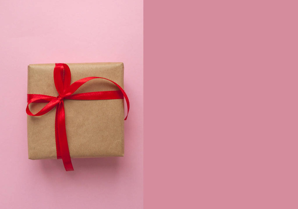 Gift box wrapped in brown colored craft paper and tied with red bow on pink background with copyspace. - Photo, Image