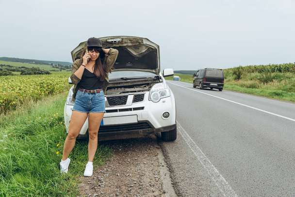 молодая взрослая женщина, стоящая рядом со сломанной машиной на шоссе и разговаривающая по телефону. Call car service
 - Фото, изображение