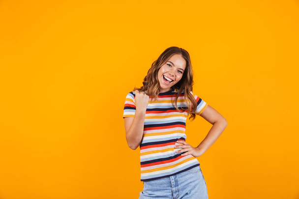 Portrait of a cheerful young girl standing isolated over yellow background, celebrating success - Фото, изображение