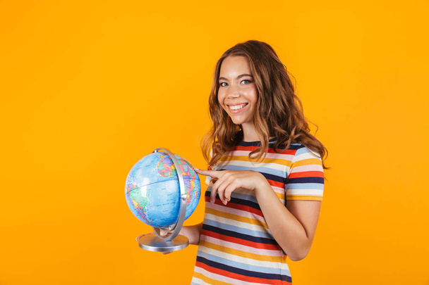 Portrait of a pretty young girl standing isolated over yellow background, pointing finger at a globe - Foto, Imagem