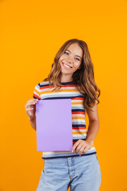Image of happy young beautiful girl posing isolated over yellow background holding blank. - 写真・画像