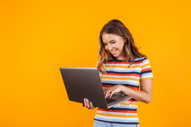Portrait of a smiling cheerful young girl standing isolated over yellow background, using laptop - 写真・画像