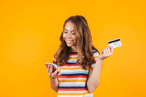 Portrait of a smiling cheerful young girl standing isolated over yellow background, showing plastic credit card, using mobile phone - 写真・画像