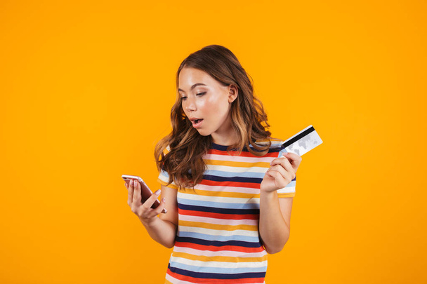 Portrait of a smiling cheerful young girl standing isolated over yellow background, showing plastic credit card, using mobile phone - Photo, Image