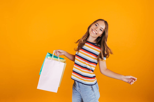 Image of a happy young girl posing over yellow wall background holding shopping bags. - Фото, изображение