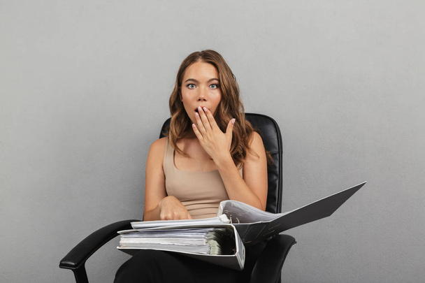 Portrait of a shocked young business woman isolated over gray background, sitting in a office chair, holding folder with documents - Фото, зображення