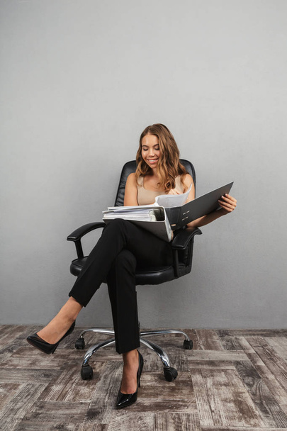 Portrait of a confident young business woman over gray background, sitting in a office chair, holding folder with documents - Fotó, kép