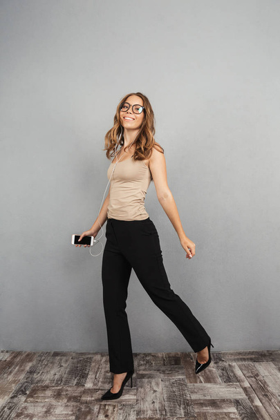 Full length portrait of a confident young business woman over gray background, listening to music with earphones and mobile phone, walking - Foto, Imagem