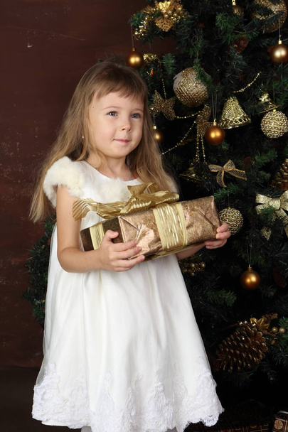A girl in a white dress near the Christmas tree holds a gift - Zdjęcie, obraz