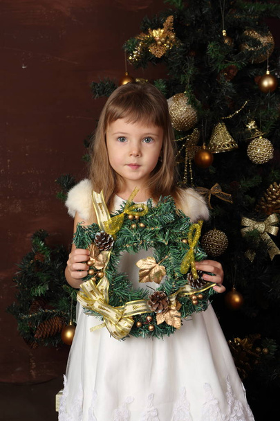 A girl in a white dress near the Christmas tree holds a gift - Φωτογραφία, εικόνα