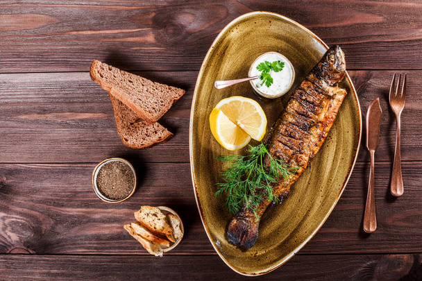 Запечённая рыба с лимоном и зеленью на деревянном фоне. Вкусное блюдо из морепродуктов. Вид сверху
 - Фото, изображение