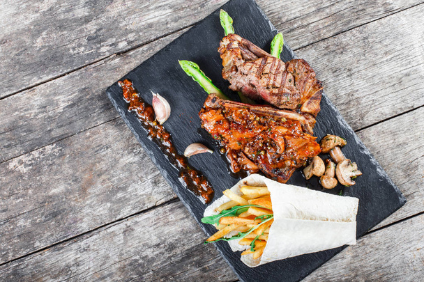 Grilled pork steaks with herbs, asparagus, garlic, mushrooms and potato fry on stone slate background on wooden background close up. Hot Meat Dishes. Top view, flat lay - Photo, Image