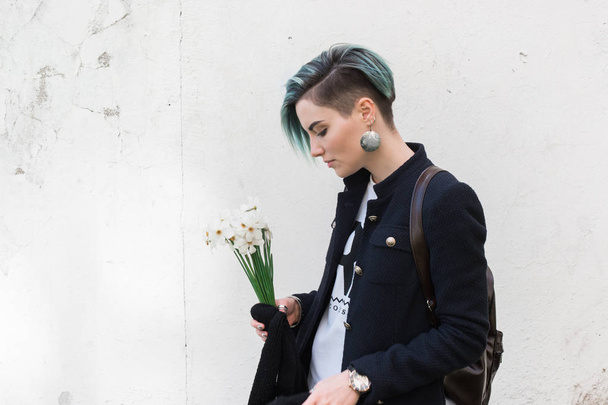 Hipster menina vestindo jaket, posando contra a parede de rua áspera com flores em suas mãos. Estilo de roupa urbana minimalista
. - Foto, Imagem
