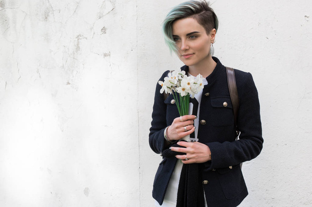 Csípő lány visel jaket, pózol elleni durva utcai fal, virág a kezében. Minimalista városi öltözködési stílus. - Fotó, kép