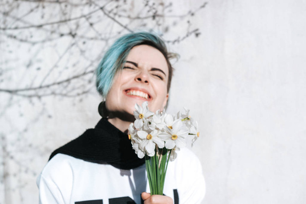 Chica Hipster posando contra la pared de la calle áspera con flores en sus manos en primavera. Sonriendo y entrecerrando los ojos del sol
. - Foto, imagen