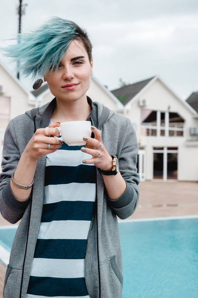 Porträt einer glücklichen Frau mit blauen Haaren, die Tee oder Kaffee in der Nähe des Pools auf dem Hinterhof ihres Ferienhauses trinkt - Foto, Bild