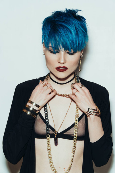 fashion photography of a girl with blue hair on a white background looking straight into the camera. Thick yellow chain and bijouterieher on her neck. Professional model posing in studio - Foto, Imagem