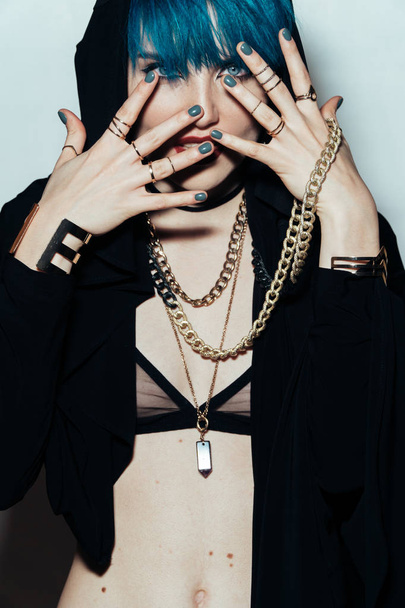 fashion photography of a girl with blue hair on a white background looking directly at the camera covers her face with her hands . Thick yellow chain and bijouterieher on her neck. - Valokuva, kuva