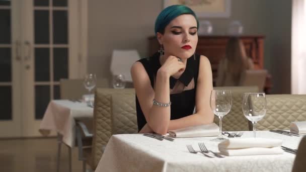 Mooie triest vrouw met kort blauw haar, alleen zittend in restaurant, wachten op iemand. Sad en depressie, lege glazen op tafel. Mislukte datum. - Video