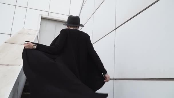 Egy fekete tunika, köpenyt, kalapot lány megy fel a lépcsőn, a háttér fehér, minimalista épület. Ő megfordul, és néz a kamerába. Lassú mozgás, lövés 120 fps. - Felvétel, videó