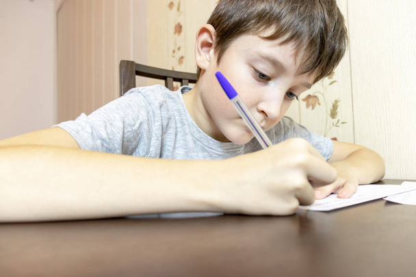 A fiú otthon az asztalnál ül, és írásban tollal papírra. - Fotó, kép