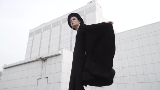 Menina em uma túnica preta, capa e chapéu se vira na frente do edifício minimalista branco. Olha em volta e acena o vestido. Slow motion shot 120 fps
. - Filmagem, Vídeo