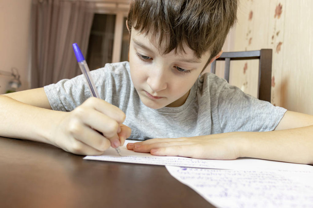 Een jongen door de tabel thuis zitten en schrijven met een pen op papier. - Foto, afbeelding