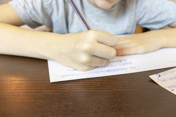 A boy sitting by the table at home and writing with a pen on paper. - Foto, imagen