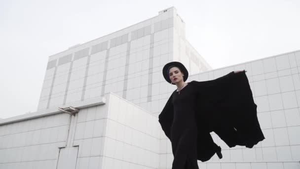 Girl in a black tunic, cloak and hat turns around in front of white minimalistic building. Looks around and waving her dress. Slow motion shot 120 fps. - Footage, Video