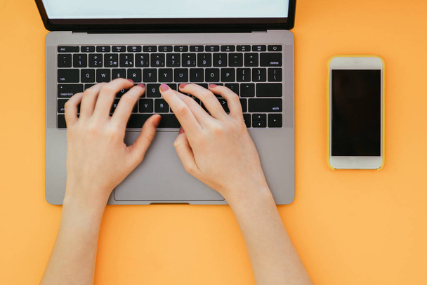 Les mains des femmes tapent le texte sur le clavier de l'ordinateur portable sur le fond orange, la vue du dessus. Travailler sur un ordinateur portable et smartphone est isolé un fond jaune
 - Photo, image