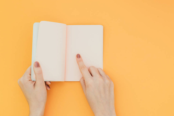 Hands hold an open notebook on an orange background, showing an index finger to a clean page, Copyspace. Flat lay - Zdjęcie, obraz