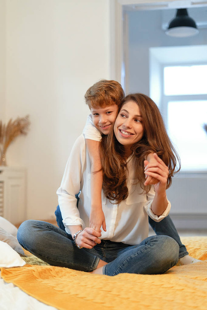 Mamá e hijo sonriendo, abrazándose, divirtiéndose, pasando tiempo juntos en un ambiente relajante en casa mientras están sentados
 - Foto, Imagen